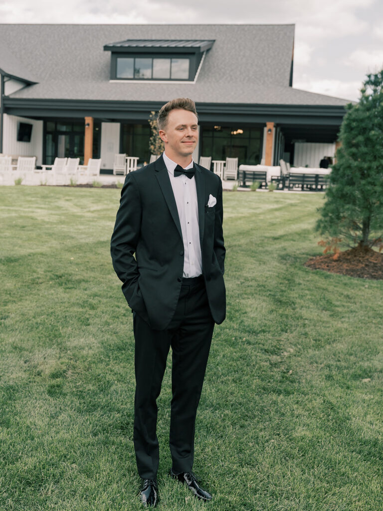 Groom in black tuxedo in front of the white farmhouse at Venue Bella Giornata